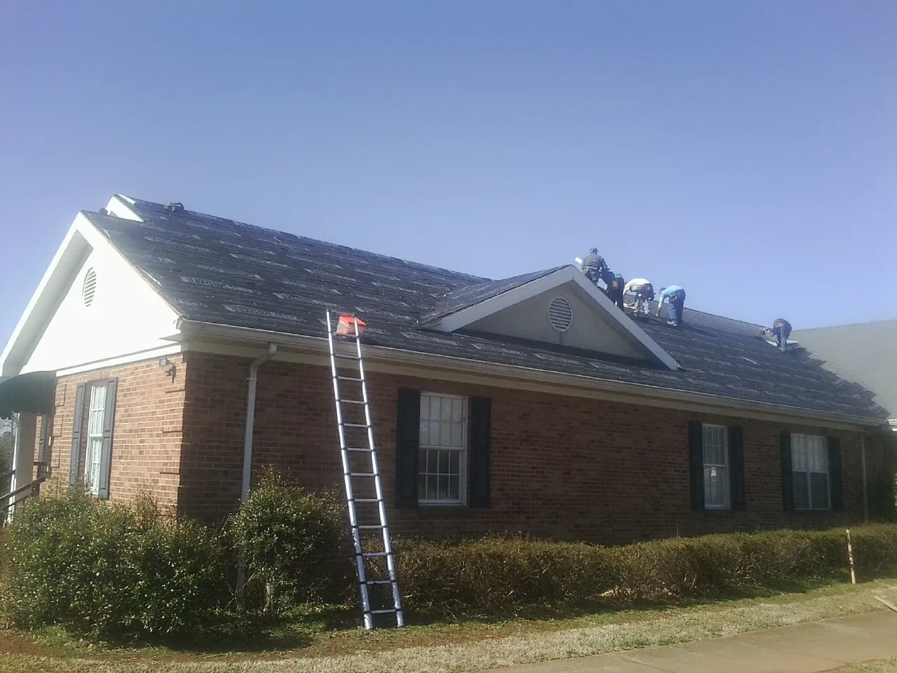 Crew tearing shingles off a brick ranch roof with a ladder set — Emergency Roof Repair work by our crew in Cedar Bluffs
