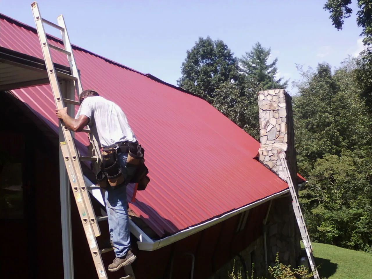Roofer on a ladder beside a red metal roof and stone chimney — Soffit & Fascia Repair by Roofing Brothers in Cedar Bluffs